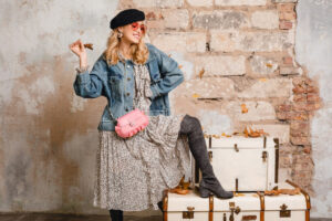 A stylish woman poses with one leg on stacked vintage trunks, wearing a denim jacket and floral dress, holding a leaf and a pink handbag.