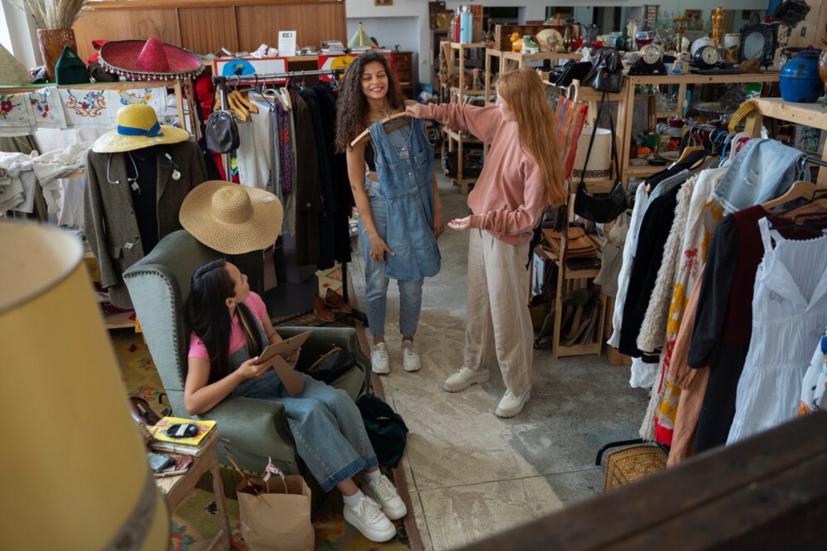 Three women are shopping in a cozy thrift store filled with colorful clothes, accessories, and decor items. One woman is trying on a dress.