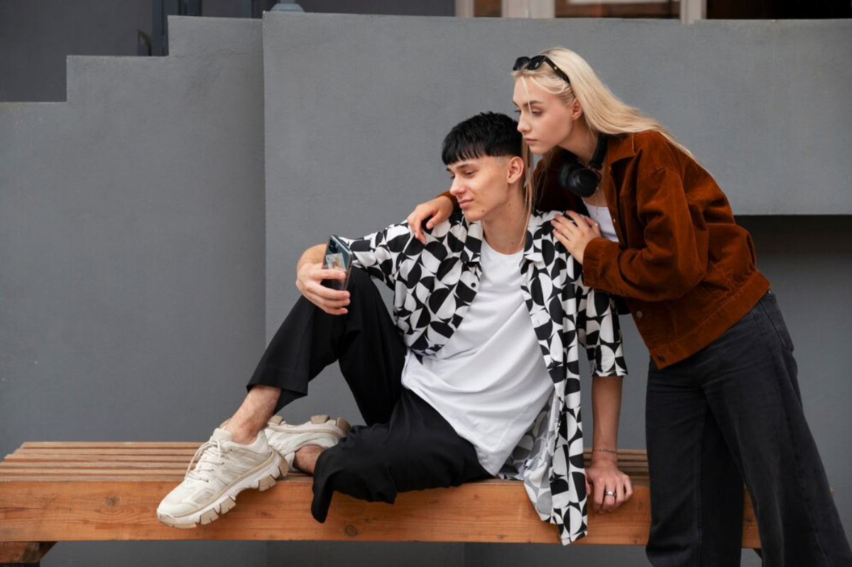 A person sits on a wooden bench in a stylish shirt, while another leans in close, sharing a playful moment against a gray backdrop.