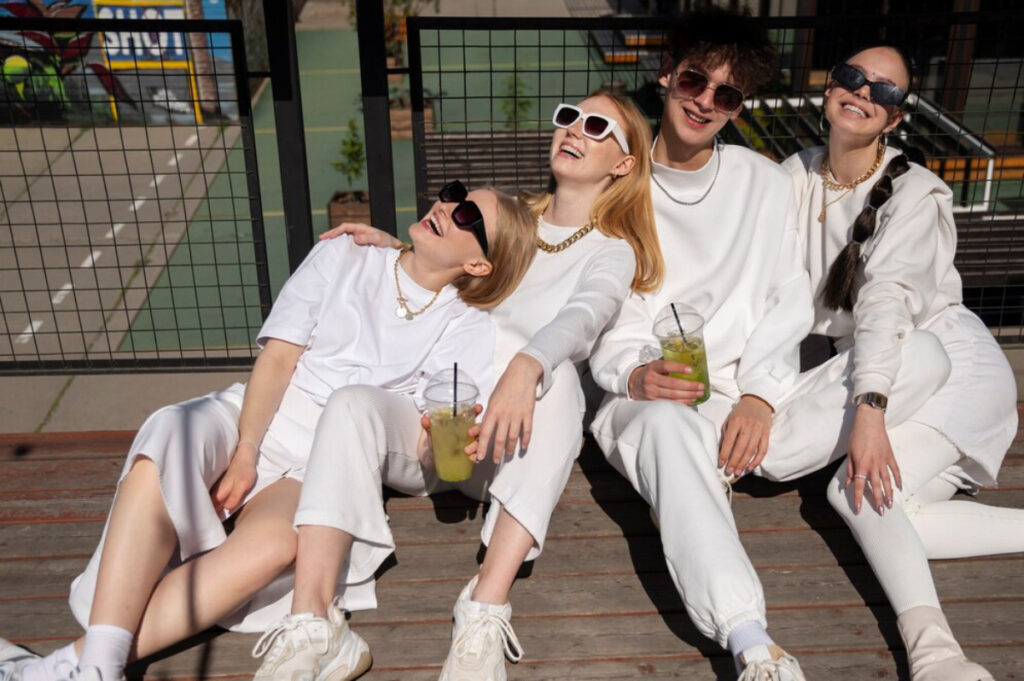 Four friends dressed in stylish white outfits, enjoying drinks while lounging on a wooden deck in a sunny outdoor setting.