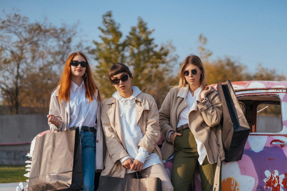 Three stylish women stand beside a colorful vintage truck, holding shopping bags, in an outdoor setting with trees in the background.