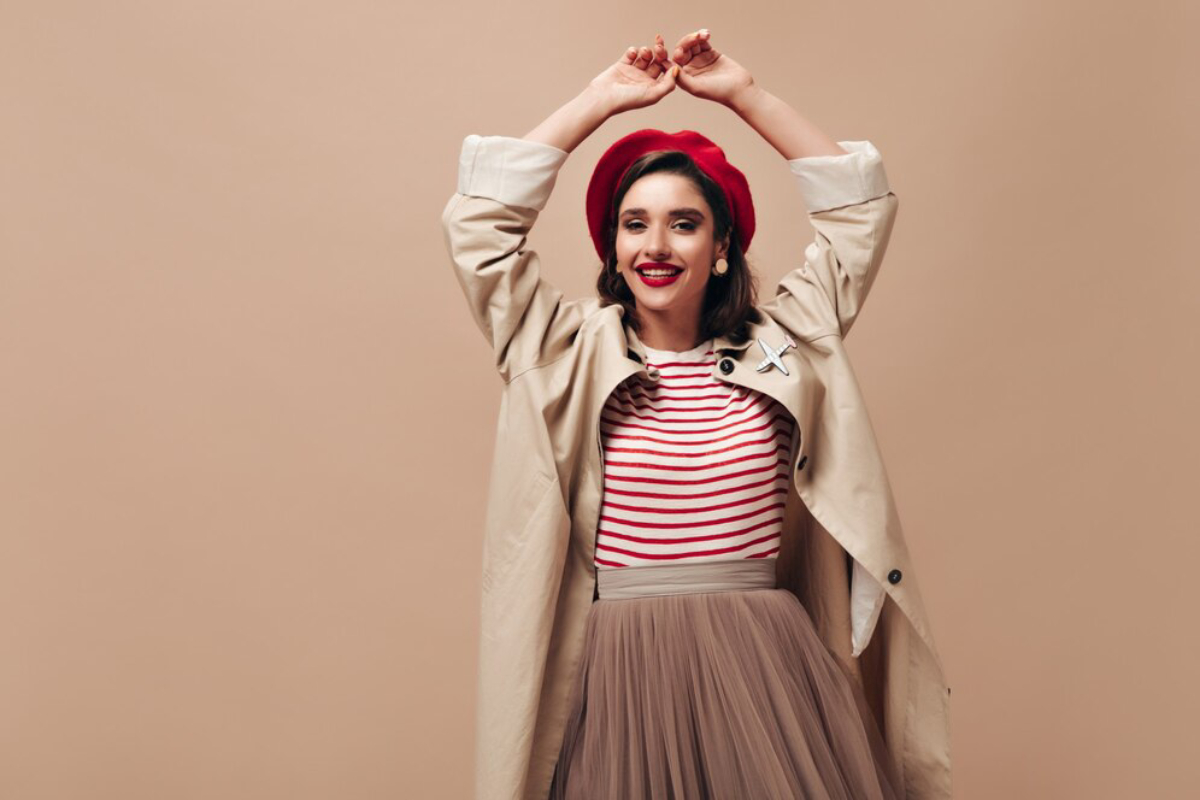 A woman poses with arms raised, wearing a red beret, striped shirt, and a beige trench coat over a flowing tulle skirt against a neutral backdrop.