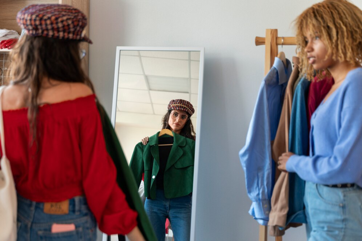 A woman in a red off-shoulder top examines herself in a mirror while holding a green jacket, surrounded by colorful clothing on hangers.