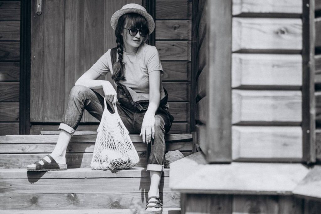Woman in sunglasses and straw hat sitting on wooden steps, holding a mesh bag, dressed in casual attire.