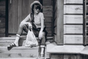 Woman in sunglasses and straw hat sitting on wooden steps, holding a mesh bag, dressed in casual attire.