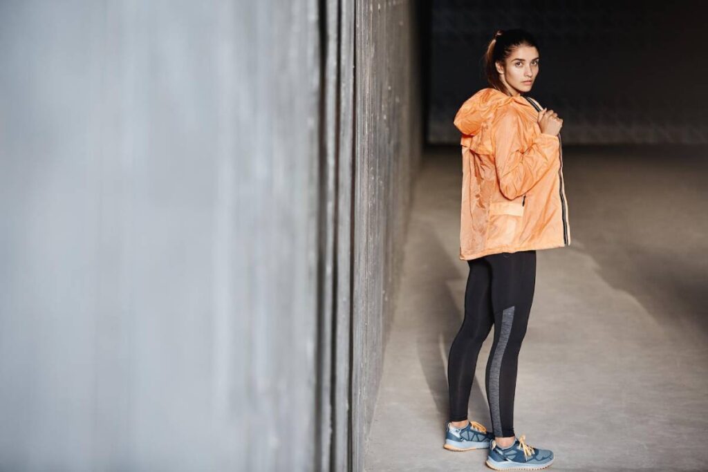 Woman in orange jacket and black leggings standing in a dimly lit hallway, looking over her shoulder.