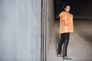 Woman in orange jacket and black leggings standing in a dimly lit hallway, looking over her shoulder.