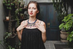 A woman in a black dress and a colorful necklace stands in a lush courtyard.