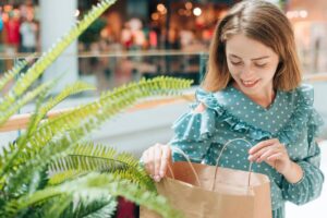 Woman in a polka dot dress smiling while looking into a shopping bag at a mall.