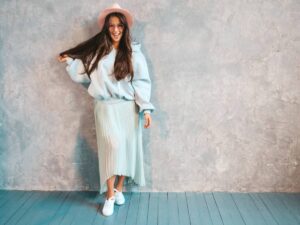 Woman in a pastel outfit and hat smiling, standing against a textured grey wall with a blue floor.