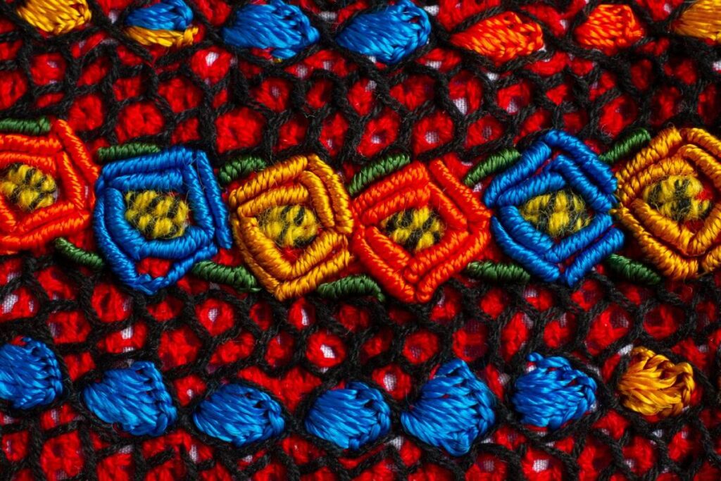 Colourful embroidered fabric featuring detailed, geometric patterns in vibrant red, blue, yellow, and green threads.