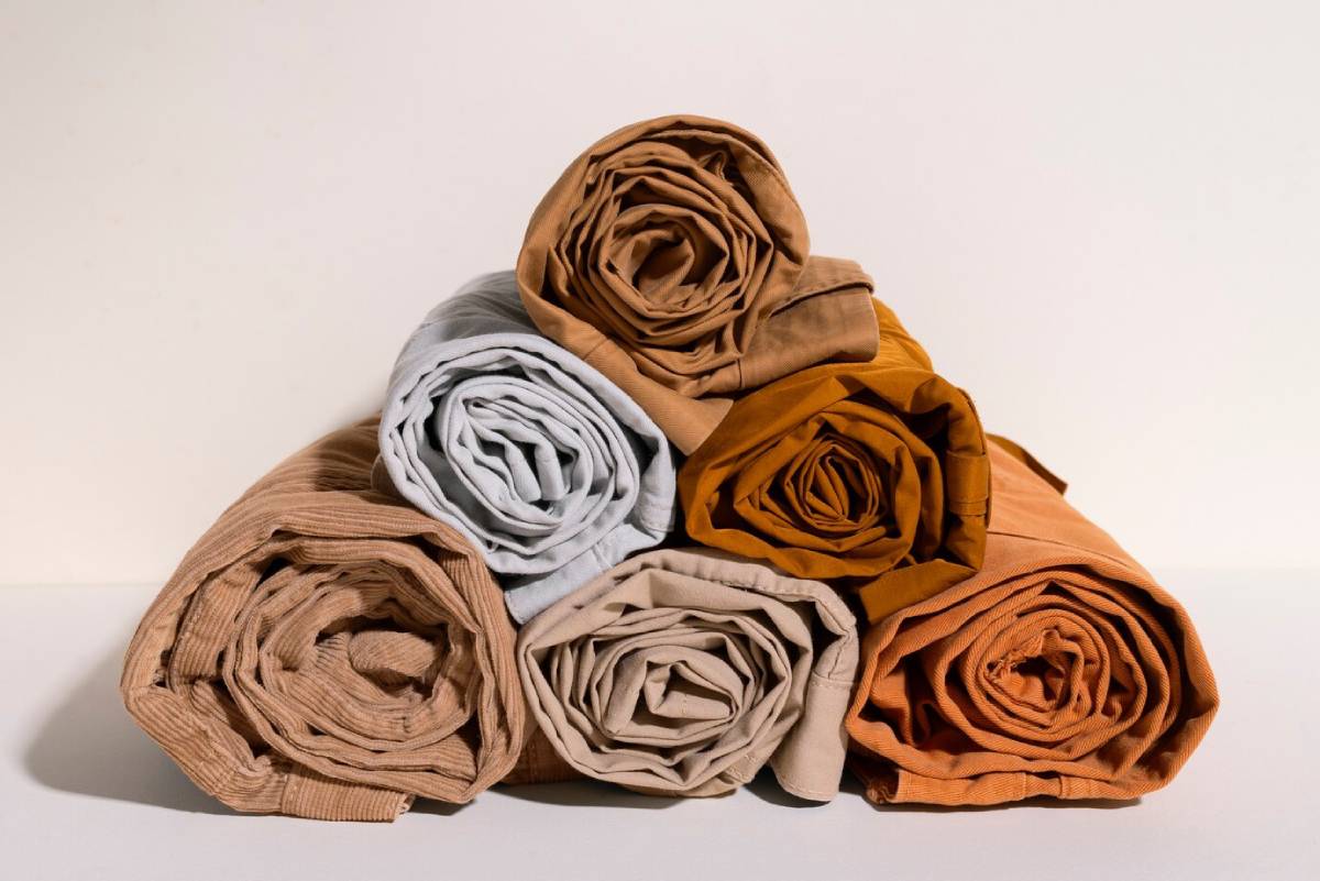 Rolled fabrics in beige, grey, and shades of brown arranged in a stack on a light background. 
