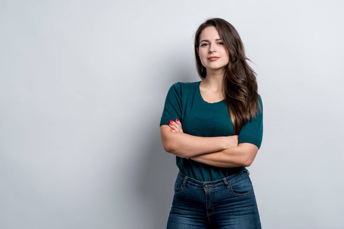 Woman in green T-shirt and jeans standing with arms crossed, smiling subtly, against a grey background.