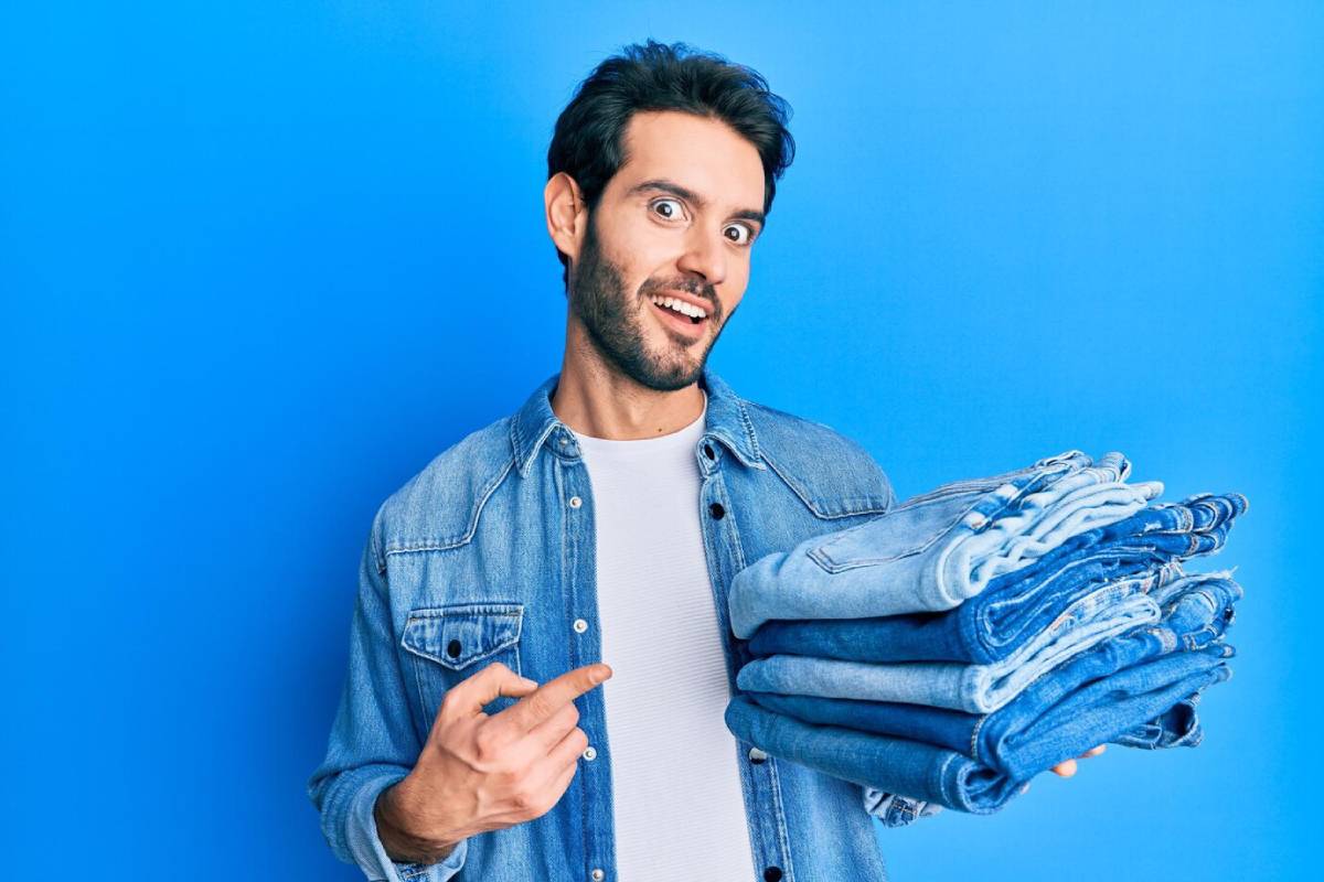  A surprised young man in a denim jacket holding a stack of jeans, against a bright blue background.