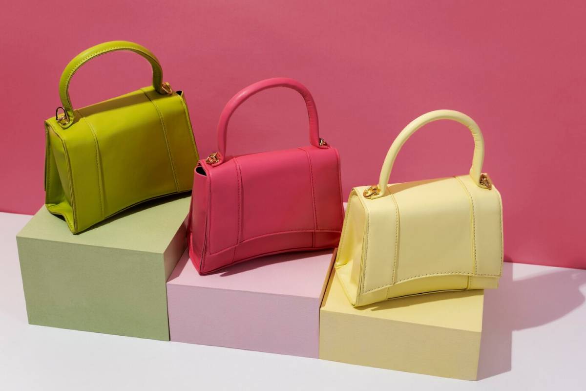 Three colourful handbags—lime green, bright pink, pale yellow—displayed on matching pastel blocks against a pink background. 
