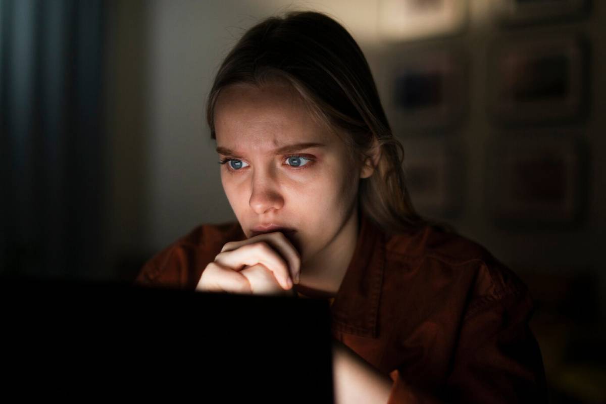 Young woman looking intently at a computer screen in a dimly lit room, appearing thoughtful and slightly worried. 