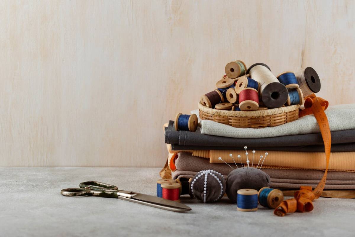 Stacked fabrics, spools of thread in a basket, scissors, and a tape measure on a neutral background, suggesting a sewing theme. 