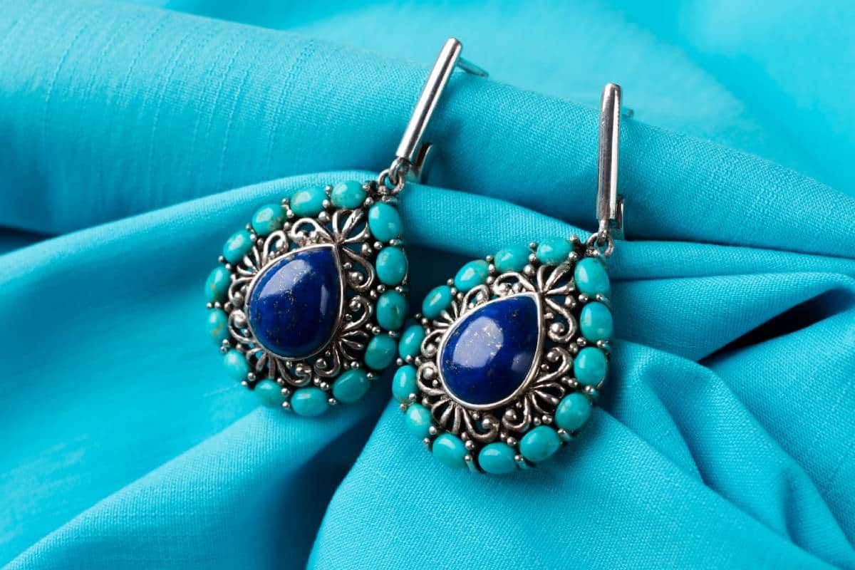 Silver filigree earrings with large blue and small turquoise stones on a teal fabric background.