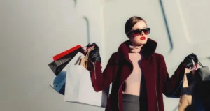 A shopper dressed in a maroon coat holds multiple colorful shopping bags against a light gray background.